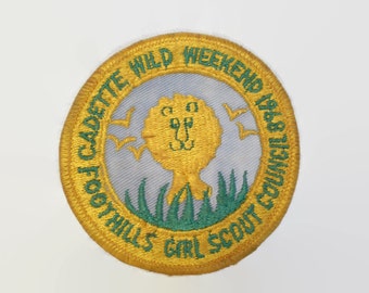 May include: A circular embroidered patch with a light blue center featuring a gold lion head, green grass, and gold birds. The gold border contains the green text CADETTE WILD WEEKEND 1968 FOOTHILLS GIRL SCOUT COUNCIL in a circular arrangement.