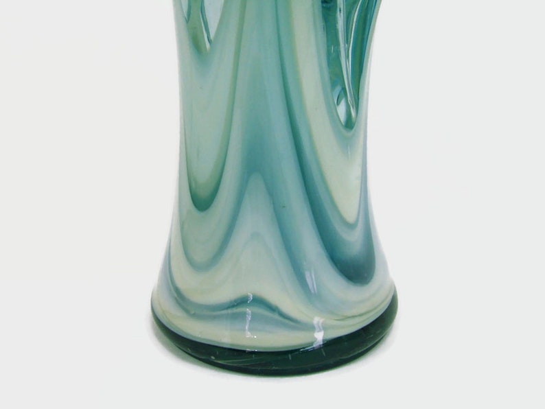 May include: A glass vase featuring a swirling pattern of translucent teal, light blue, and opaque white colors. The design consists of vertical, elongated streaks that blend together, creating a fluid, marbled appearance across the surface.