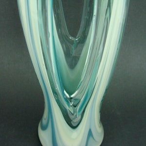 May include: A tall glass vase featuring a swirling pattern of translucent teal, light blue, and opaque white colors. The glass has a smooth, polished surface with a V-shaped opening at the top and a flared base, showcasing fluid, elongated vertical lines.