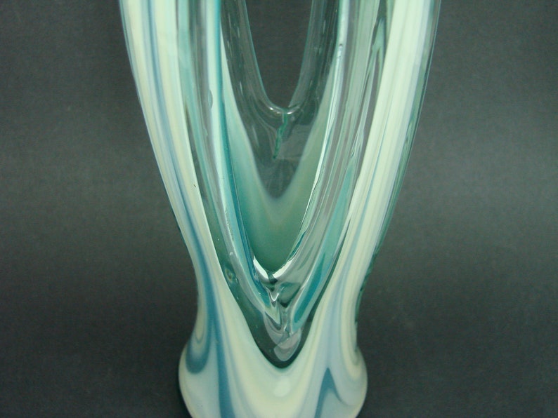 May include: A tall glass vase featuring a swirling pattern of translucent teal, light blue, and opaque white colors. The glass has a smooth, polished surface with a V-shaped opening at the top and a flared base, showcasing fluid, elongated vertical lines.