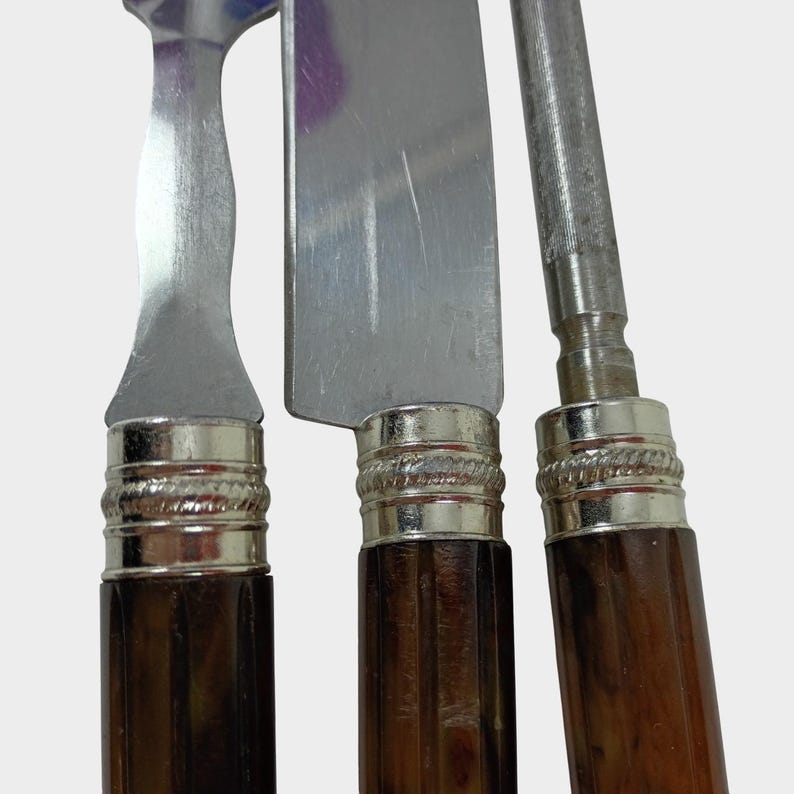 Puede incluir: Primer plano de tres cuchillos de acero inoxidable con mangos de madera marr&oacute;n oscuro. Los mangos est&aacute;n acentuados con bandas de metal plateado. Las hojas son brillantes y reflejan la luz. Los cuchillos parecen ser parte de un juego de cubiertos.
