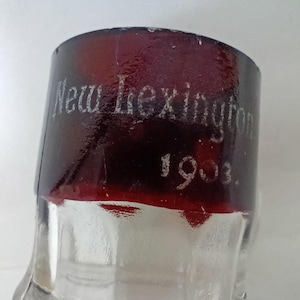 May include: Close-up of a clear glass bottle with a dark red band at the top. The band has the words "New Lexington" and the year "1903" etched in white.