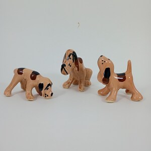 May include: Three ceramic dog figurines in various poses. The dogs are tan with brown spots and black accents. One dog is sniffing the ground, another is sitting, and the third is standing.