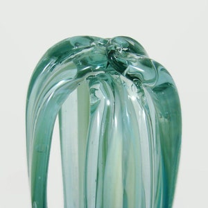 May include: A translucent light blue glass sculpture featuring elongated, curved ribbons of glass that converge at the top. The object has a smooth, glossy finish with subtle variations in color intensity throughout the vertical glass strands.