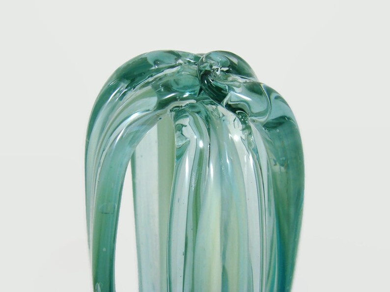 May include: A translucent light blue glass sculpture featuring elongated, curved ribbons of glass that converge at the top. The object has a smooth, glossy finish with subtle variations in color intensity throughout the vertical glass strands.