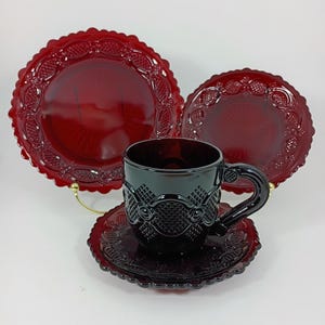 May include: Ruby red glass teacup and saucer set with matching plates. The cup and saucer have a detailed pattern, and the plates have a scalloped edge. The set is displayed on a white background.
