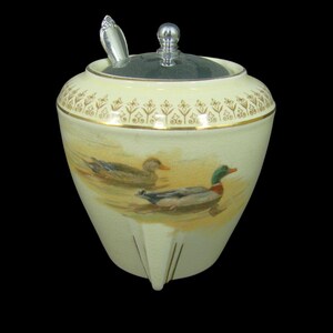 May include: A white ceramic honey pot with a silver lid and a silver spoon. The pot has a gold trim and a design of two ducks on the front.