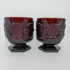 May include: Two ruby red goblets with intricate diamond and floral patterns. The glasses have a square base and a rounded bowl. The design features a series of diamond shapes and floral accents, creating a vintage aesthetic. The goblets are made of glass.