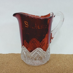 May include: A ruby red glass pitcher with a clear handle and a decorative, cut-glass base. The pitcher has the word "Salerno" etched in gold lettering. The pitcher is likely vintage and suitable for serving beverages.