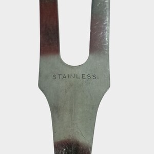 Puede incluir: Primer plano de un utensilio de cocina de acero inoxidable. La palabra "STAINLESS" est&aacute; grabada en el metal. El utensilio tiene un dise&ntilde;o de dos puntas con un mango curvo.