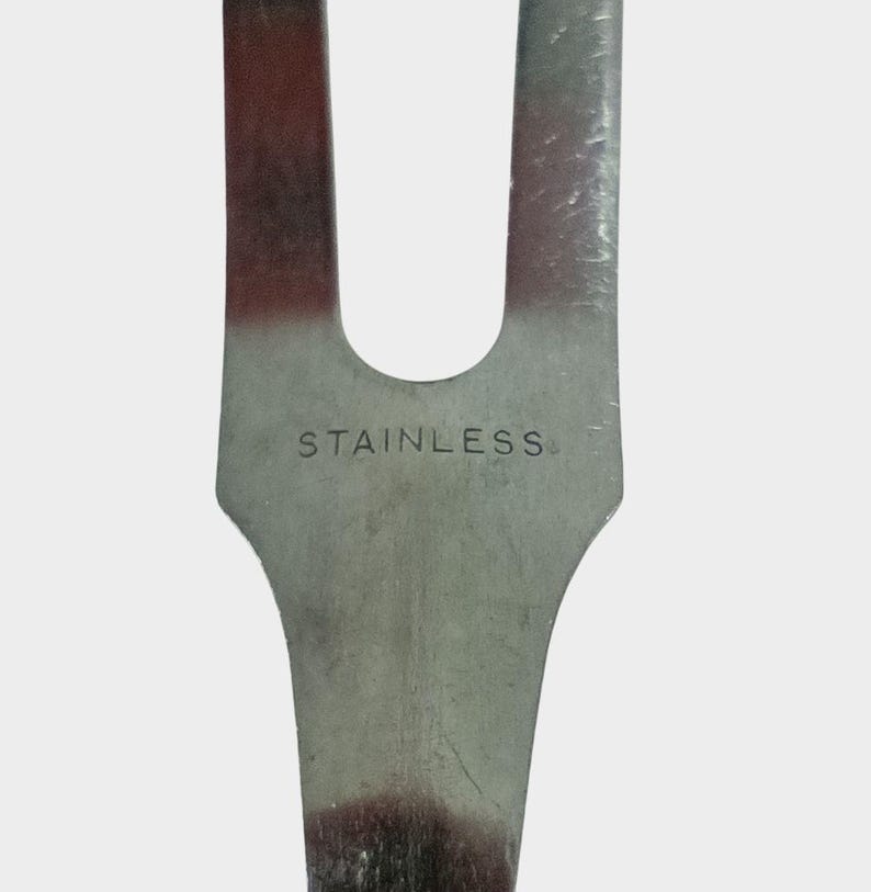 Puede incluir: Primer plano de un utensilio de cocina de acero inoxidable. La palabra "STAINLESS" est&aacute; grabada en el metal. El utensilio tiene un dise&ntilde;o de dos puntas con un mango curvo.