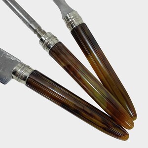 Puede incluir: Primer plano de tres cuchillos antiguos. Las hojas y los detalles son de color plateado, mientras que los mangos son de color marr&oacute;n c&aacute;lido con un efecto veteado. Los cuchillos est&aacute;n dispuestos en diagonal, mostrando su dise&ntilde;o y artesan&iacute;a.