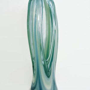 May include: A tall, slender glass vase featuring a swirling pattern of translucent teal, light blue, and white colors. The cylindrical body has a smooth, polished surface with vertical striations that taper slightly toward the rounded top opening.