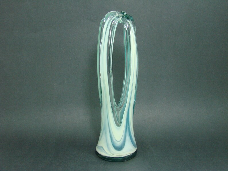 May include: A tall, clear glass vase featuring a swirling pattern of opaque white and translucent teal blue glass. The vase has a wide, circular base that tapers upward into two distinct, elongated vertical loops that meet at the top.