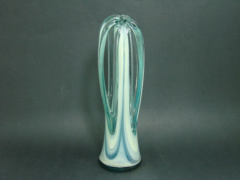 May include: A tall, slender glass vase featuring a swirling pattern of translucent teal and opaque white glass. The design includes elongated, arched glass loops that extend upward from the base, creating an open, airy structure with a smooth finish.