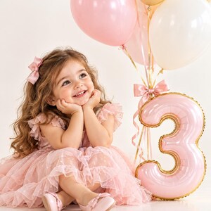 May include: A child with long wavy hair wearing a pink ruffled tulle dress and matching bow hair clip, sitting beside a large pink foil balloon shaped like the number three, accompanied by a cluster of pink, white, and gold helium balloons.