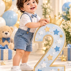 May include: A large, light blue number two prop decorated with gold and white stars and topped with a gold crown. The number features a smooth, marbled finish and gold-toned edges, designed as a decorative piece for a second birthday celebration.