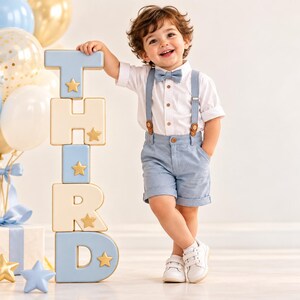 May include: A white button-down shirt, light blue shorts with matching suspenders, and a light blue bow tie. A stack of block letters spelling THIRD in light blue and cream with gold star accents stands beside the outfit, with balloons.