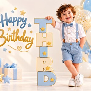 May include: A toddler wearing a white button-down shirt, a light blue bowtie, light blue suspenders, and matching shorts, standing next to a vertical stack of light blue and cream-colored blocks spelling the word THIRD with gold star accents.