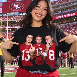 May include: A black short-sleeved t-shirt featuring a graphic print of three football players in red jerseys with the numbers 13, 16, and 8. The central player holds a beer bottle, while the others hold cigarettes and make hand gestures.
