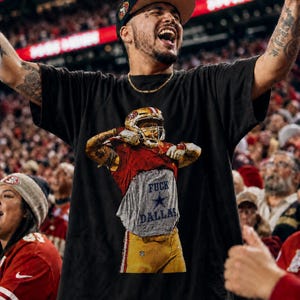 May include: A black t-shirt featuring a graphic of a football player in a red and gold uniform lifting their jersey to reveal a gray undershirt with the text FUCK DALLAS and a blue star logo printed on the front of the garment.