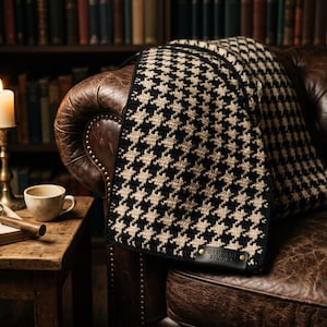 May include: A black and cream houndstooth patterned blanket featuring a dark leather tag embossed with the text Obsidian Studio, secured with two metal rivets at the bottom corner. The textile has a textured, woven appearance with a repeating pattern.