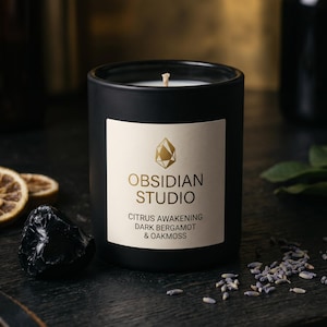 May include: A matte black candle jar with a white label featuring the gold text Obsidian Studio, Citrus Awakening, Dark Bergamot & Oakmoss. The candle has a single centered wick and is surrounded by dried citrus slices and lavender.