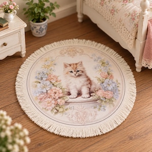 Può includere: Un tappetino tessile rotondo color crema con un'illustrazione centrale di un soffice gattino marrone chiaro seduto su un piedistallo. Il gattino è circondato da una ghirlanda di fiori rosa e blu, rifinita con un bordo decorativo a frange.