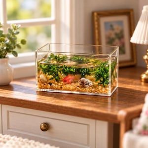 May include: A rectangular clear acrylic aquarium containing water, green aquatic plants, tan gravel, two small seashells, and a miniature orange goldfish figurine. The transparent tank is positioned on a wooden surface, showcasing the internal contents.