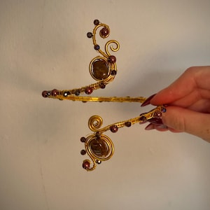 May include: An arm cuff crafted from gold-toned wire, featuring a spiral design at each end. The piece is accented with dark brown, polished stones and small, round, deep maroon beads that are wrapped along the wire structure.