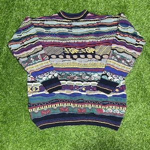 Puede incluir: Un suéter de manga larga con cuello redondo que presenta un complejo patrón de punto multicolor con bandas horizontales en azul, rojo, amarillo, verde y negro. La prenda muestra un tejido texturizado en relieve con diversas formas geométricas.