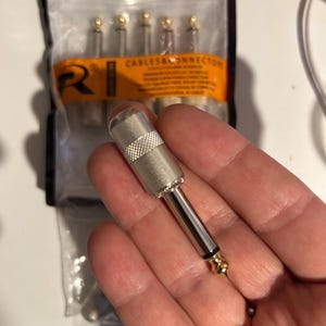 May include: A silver-colored 1/4 inch audio jack connector with a knurled grip section and a gold-plated tip. In the background, a clear plastic storage bag contains additional identical audio connectors with an orange label reading CABLES&ONNECTORS.