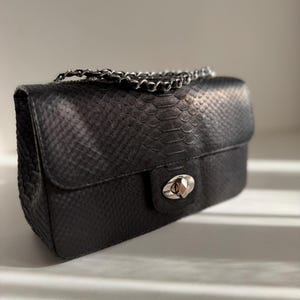 May include: A black rectangular handbag with a textured snakeskin pattern. The bag features a silver-toned metal turn-lock closure on the front flap and a matching silver-toned metal chain strap resting on top of the bag.