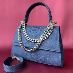 May include: A blue handbag with a textured snakeskin pattern, featuring a single top handle, a silver-toned metal chain draped across the front flap, and a matching blue snakeskin-textured wrist strap positioned to the left of the bag.