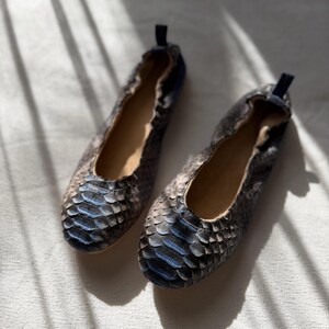 Könnte beinhalten: Ein Paar Slipper-Ballerinas mit einem strukturierten, schuppenartigen Muster in Dunkelgrau, Schwarz und schillerndem Blau. Die Schuhe haben eine abgerundete Spitze, einen gerafften, elastischen Rand und eine kleine dunkle Zuglasche an der Ferse.