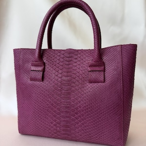 Könnte beinhalten: Eine tiefviolette Handtasche mit einem strukturierten Schlangenhautmuster auf der gesamten Außenseite. Die Tasche verfügt über zwei abgerundete Tragegriffe, die mit rechteckigen Laschen befestigt sind, und einen strukturierten Körper.