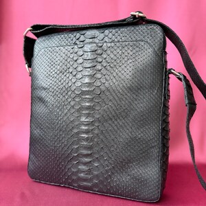May include: A black crossbody bag featuring a textured snakeskin pattern across the front panel. The bag has a square silhouette, a top zipper closure, and an adjustable shoulder strap attached with silver-toned metal hardware rings.