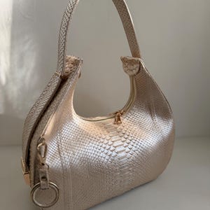 Puede incluir: Un bolso de mano en forma de media luna en un tono dorado champán brillante con un patrón texturizado de piel de serpiente. El bolso cuenta con un asa superior, un compartimento principal con cremallera y un anillo metálico en el lateral.