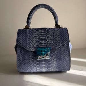 May include: A structured handbag featuring a textured, blue snakeskin-patterned exterior. The bag has a single top handle, a flap closure, and a metallic silver-toned clasp mechanism at the center, which is partially covered by a protective blue film.