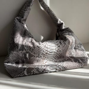 May include: A hobo-style shoulder bag featuring a textured grey and white snakeskin pattern. The bag has a slouchy, unstructured silhouette with a single integrated shoulder strap and a wide, curved opening that tapers toward the top handle.