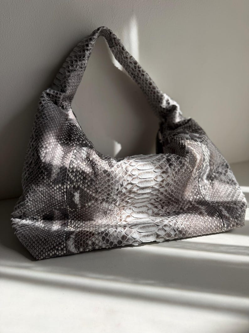May include: A hobo-style shoulder bag featuring a textured grey and white snakeskin pattern. The bag has a slouchy, unstructured silhouette with a single integrated shoulder strap and a wide, curved opening that tapers toward the top handle.