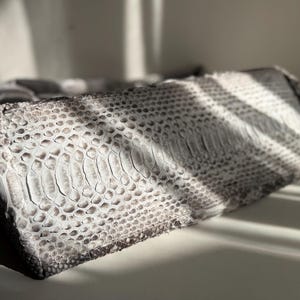 May include: A rectangular clutch bag featuring a textured snakeskin pattern in shades of grey, white, and black. The surface displays a distinct scale-like appearance with a gradient of light and dark tones across the exterior of the accessory.