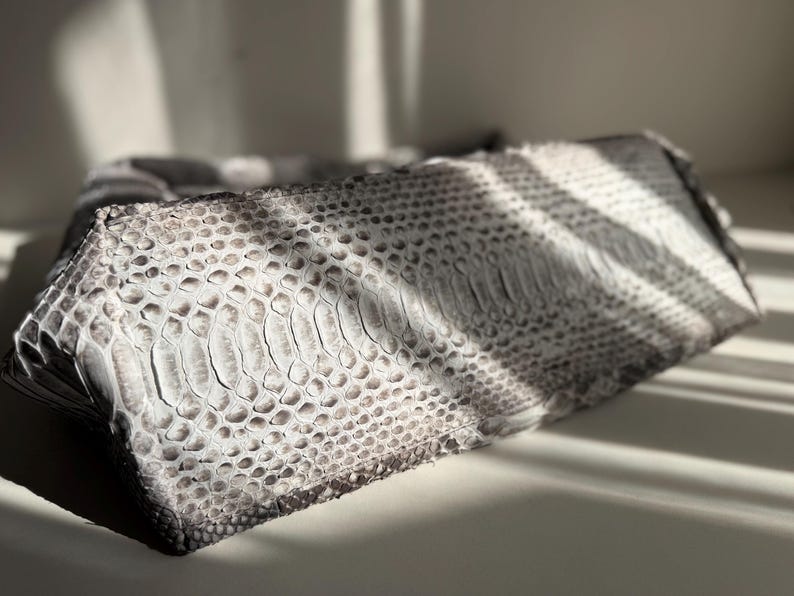 May include: A rectangular clutch bag featuring a textured snakeskin pattern in shades of grey, white, and black. The surface displays a distinct scale-like appearance with a gradient of light and dark tones across the exterior of the accessory.