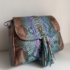 May include: A crossbody bag featuring a snakeskin-patterned texture in metallic copper, iridescent blue, and purple tones. The bag has a fold-over flap secured with a silver-toned metal clasp and a matching leather tassel hanging from the front.