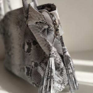 May include: A handbag featuring a grey and white snakeskin pattern with a textured surface. The bag includes a silver metal zipper closure at the top and two matching snakeskin-patterned tassels hanging from the zipper pull, adding detail.