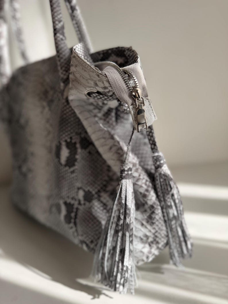 May include: A handbag featuring a grey and white snakeskin pattern with a textured surface. The bag includes a silver metal zipper closure at the top and two matching snakeskin-patterned tassels hanging from the zipper pull, adding detail.