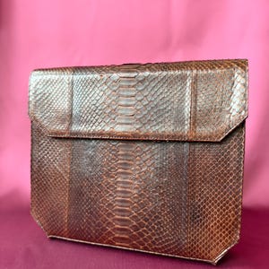 Könnte beinhalten: Eine rechteckige braune Clutch mit strukturiertem Schlangenledermuster. Die Tasche verfügt über einen strukturierten Klappverschluss, der sich über die Oberseite erstreckt, mit einem durchgehenden Schuppenmuster auf der Oberfläche und abgeschrägten Ecken.