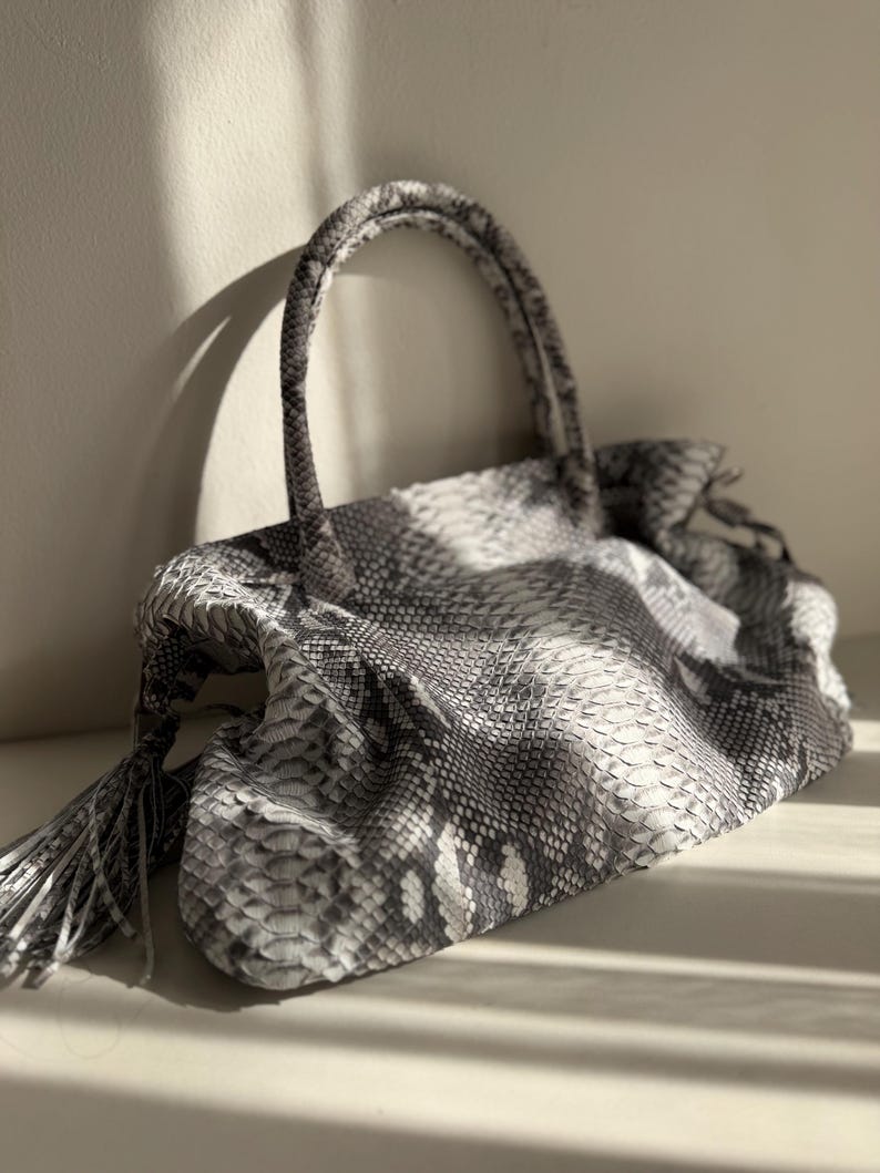 May include: A handbag featuring a grey and white snakeskin pattern with a textured scale finish. The bag has two rounded top handles and a decorative tassel detail hanging from the side, showcasing a slouchy, unstructured shape.
