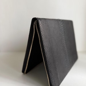 May include: A black clutch bag with a textured snakeskin pattern. The bag is folded in a triangular shape, revealing two silver-toned metal zippers that run along the edges. The surface features a repeating scale-like texture throughout the exterior.