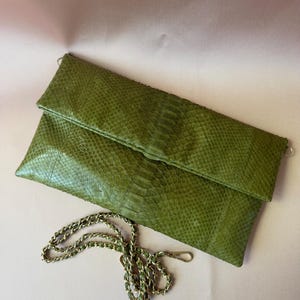 May include: A rectangular olive green clutch bag featuring a textured snakeskin pattern. The bag has a fold-over flap design and includes a detachable gold-toned metal chain strap with a lobster clasp, allowing for versatile carrying options.